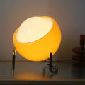 Egg-shaped glass table lamp in warm yellow glow with chrome stand on wooden shelf creating ambient lighting | Alinora