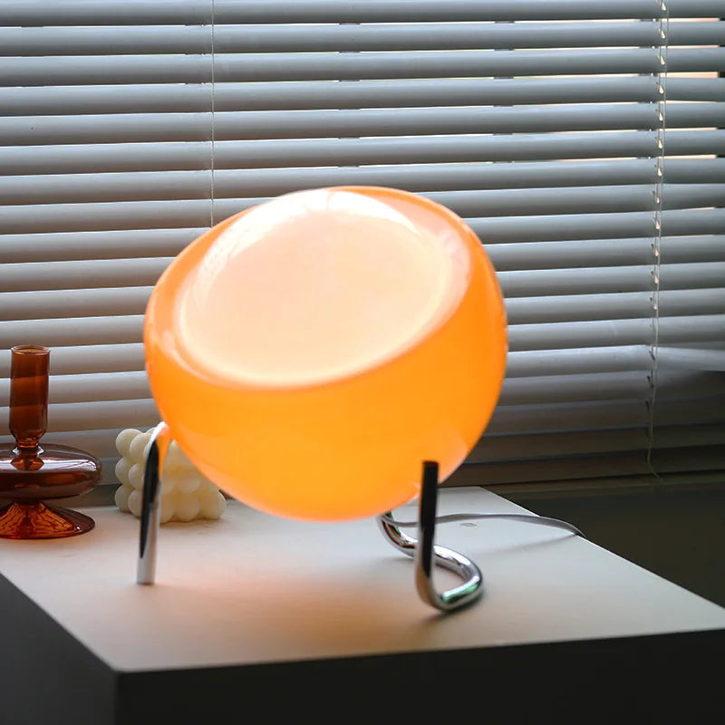 Vibrant orange egg-shaped glass table lamp with chrome stand glowing warmly on white surface against window blinds | Alinora