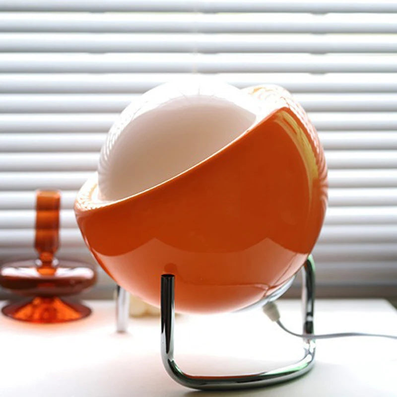 Vibrant orange egg-shaped glass table lamp with chrome stand glowing warmly on white surface against window blinds | Alinora